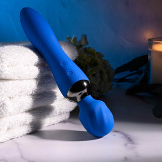 Mitt i vita handdukar och mjuk belysning vilar Selopa Blue Belle Wand, en USB-uppladdningsbar personlig massageapparat med en innovativ dubbelsidig design, nära ett tänt ljus och grönt blad för att skapa en avkopplande atmosfär.
