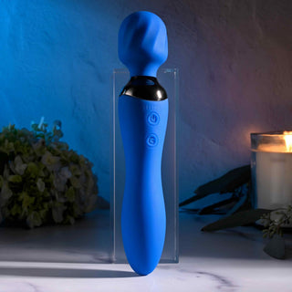 Selopa Blue Belle Wand, med ett flexibelt rundat huvud och intuitiva knappar, står upprätt mitt i mjuk belysning med ett ljus, blommor och en liten växt. Dess USB-uppladdningsbara design ger bekvämlighet för användning var som helst.