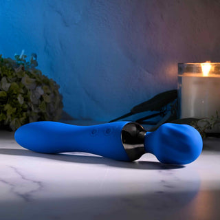 Selopa Blue Belle Wand, en USB-uppladdningsbar massageapparat med en elegant blå design, vilar elegant på en marmoryta. Mjuk belysning och ett tänt ljus förhöjer atmosfären, medan gröna bladverk i bakgrunden bidrar till den lugna och intima atmosfären.