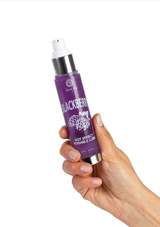 En hand som håller i en lila flaska "Secret Play Kyssbart Glidmedel" med en pumpdispenser. Flaskan är prydd med björnbärsillustrationer och silverdetaljer mot en vanlig vit bakgrund.