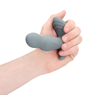 En hand håller den roterande prostatastimulatorn, med en grå, böjd, strukturerad design gjord av kroppssäker silikon mot en vit bakgrund.