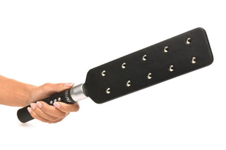 En hand som håller i E-Stim Spiked Paddle, tillverkad av elegant svart veganskt läder med metalldubbar, perfekt för att slå eller slå.