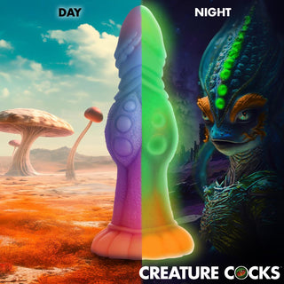 Delad bild: Day avslöjar ett färgstarkt utomjordiskt landskap; natten avslöjar en glödande varelse med levande design. Text: CREATURE COCKS, med vår Glow-In-The-Dark Alien Dildo gjord av ftalatfritt silikon för en utomjordisk upplevelse.