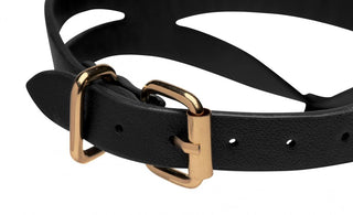Närbild av Bondage Halsband Baddie, ett svart fuskläderbälte med guldspänne och o-ring, som visar en utskuren bladdesign på en vit bakgrund.