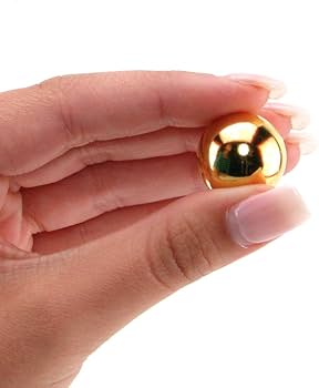 En manikyrerad hand håller en guldglänsande Klassiska Ben Wa-knipkulor mellan tummen och pekfingret, perfekt för bäckenbottengymnastik eller bäckenbottenövningar.