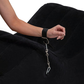 En persons arm hålls av en svart lädermanschett kopplad till en kedja fäst vid Uppblåsbaren The Lounge Sexkudde, en ergonomiskt designad svart böjd pjäs. Den vita bakgrunden framhäver installationen och betonar komfort och stil.
