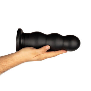 En person håller XXL Ribbad Buttplug från BUTTR, en stor svart silikondildo med en utsvängd bas, mot en vanlig bakgrund.