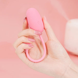 En hand håller Magic Motion Kegeltränare Rosa, en äggformad enhet med en ögla, mot en mjuk rosa bakgrund – perfekt för att boosta dina kegelövningar.