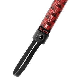 Närbild av BegMe Vegan Leather Floggers hållbara rödmönstrade handtag med en strukturerad design och svart ögla rem, med "BEGME" i svart på en röd etikett nära remmen, allt utformat för att förbättra din BDSM-upplevelse.