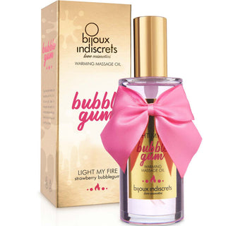 En flaska Bijoux Indiscrets Massageolja Bubblegum med en rosa rosett sitter bredvid sin guld- och rosa förpackning och erbjuder en värmande massageolja-upplevelse med bubbelgumssmak.