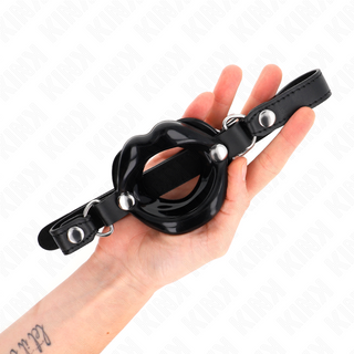 En hand håller i KINK Lip Gag 8 cm, en munkavle i svart silikon med remmar och metallnitar som kombinerar funktionalitet och stil för bondagelekar.