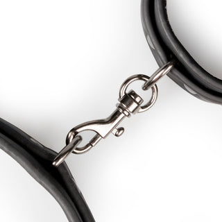 Närbild av en EasyToys Handbojor & Lårbojor vridbar karbinhake i metall som sammanfogar två svarta gummihandtag, vilket tyder på mångsidig användning i BDSM-spel mot en vit bakgrund.