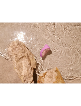 Ett rosa snäckskal på våt sand med porlande vatten bredvid två beige stenar frammanar de mjuka kurvorna och den raffinerade designen hos LELO SONA 2 Travel, som har innovativ SenSonic-teknik.