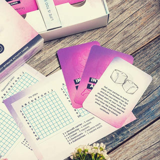 En närbild av Love Battleship Lesbisk Intimitetsspel utspelar sig på en träyta. Korten visar rosa och lila gradienter som liknar akvarellillustrationer, med ett synligt partiturblad med bokstäver och rutnät. En vit ruta är delvis synlig i bakgrunden.