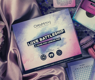En förpackad uppsättning Love Battleship Lesbisk Intimitetsspel är placerad på silkeslen rosa tyg, tillsammans med en macaron, snäckskal, en broschyr och ett rutnätskort. Boxen har pastellillustrationer. Perfekt för dejtkväll!.