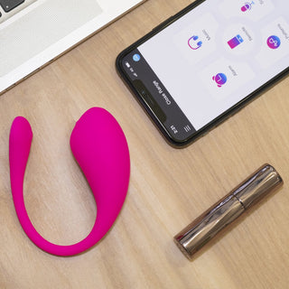 En rosa pryl bredvid en telefon som visar en app, med en läppstiftstor "Lovense Lush 3 Appstyrd Vibrator" som antyder G-punktsstimulering, är delvis inramad av en snygg bärbar dator på skrivbordet.