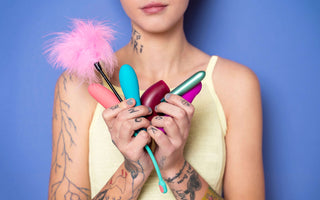En person med tatueringar håller flera sexleksaker i händerna, inklusive en rosa fjäder, färgglada vibratorer och en sugstimulator, mot en blå bakgrund.