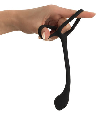 En hand håller Penis & Pungring Med Buttplug, en svart, flexibel silikon sexleksak med C-formade öglor.
