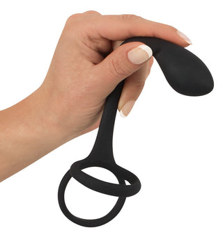 En hand håller den eleganta, svarta Penis & Pungring Med Buttplug med en böjd spets mot en vit bakgrund – ett perfekt komplement till alla silikon sexleksaker-kollektioner.
