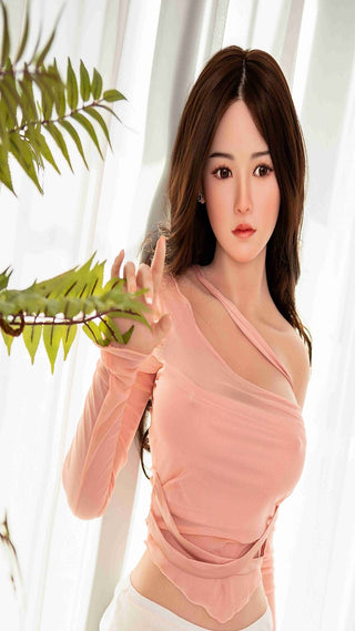 135 cm lång sexdocka i asiatisk stil av TPE med långt brunt hår och rosa topp, som står bredvid en grön växt.