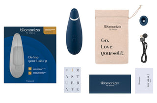 Womanizer Premium 2 i blått visas med dess förpackning, laddningskabel, en påse märkt "Go. Love yourself!", och häften. Med Pleasure Air™-teknologi för överseende, uppmanar förpackningen dig att "Definiera din lyx", eftersom #IMASTURBATE hashtaggen innebär ren tillfredsställelse.