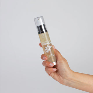 En hand håller en flaska Secret Play Kyssbart Glidmedel med pumpdispenser. Flaskan har en benvit etikett prydd med text och en illustration av en vaniljblomma, mot en vanlig ljusgrå bakgrund.