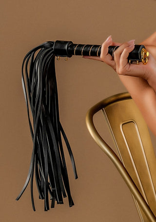 En hand tar tag i TabooM Studded Flogger med sitt svarta PU-läder-handtag, när den graciöst vilar mot en lyxig guldstol.