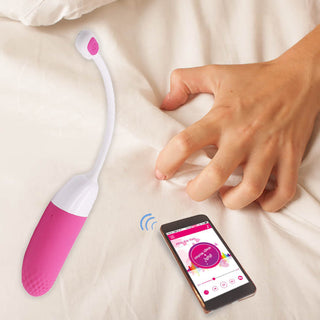 En hand sträcker sig efter en smartphone som visar en app, bredvid en Magic Motion Vini - Vibrerande Ägg, på en säng.
