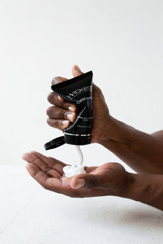 En person doserar Wicked Onani Glidmedel 120ml, ett högkvalitativt glidmedel, från en elegant svart tub i handflatan mot en orörd vit bakgrund.